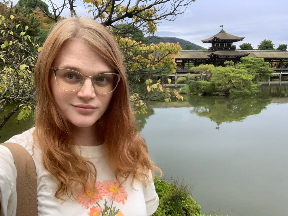 a pic of me (white girl, red hair, cat eye glasses, white top with flowers on it) in front of a pond with a japanese style building in the background