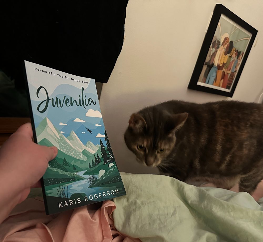 A photo of the paperback copy of JUVENILIA: POEMS OF A TWELFTH GRADE YEAR by Karis Rogerson, which is blue and green with a mountainscape, and a cat sort of sniffing the book. 