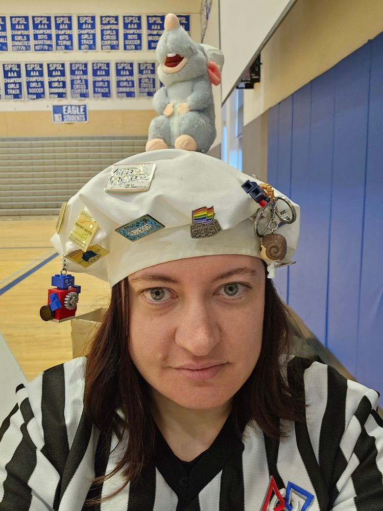 A white lady in a black/white referee stripes shirt. She has a white chef hat covered in FLL buttons and Lego Superhero Keychain. There is a Remy mouse on the very top.