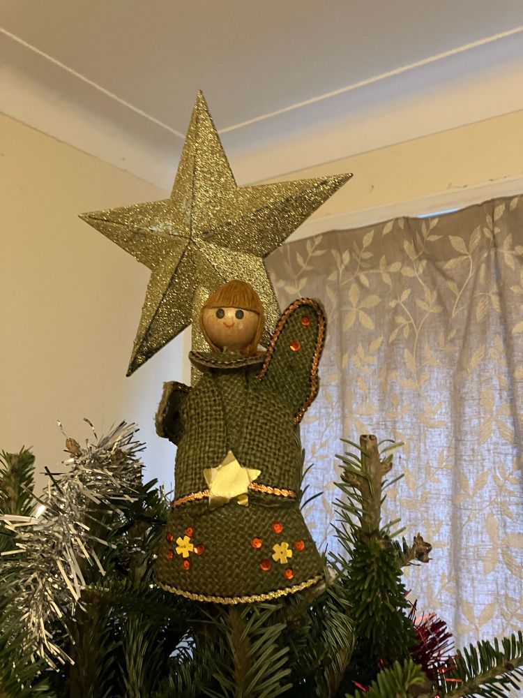 The top of a Christmas tree with a star and a battered green angel made of canvas, cardboard and wood holding a star.
