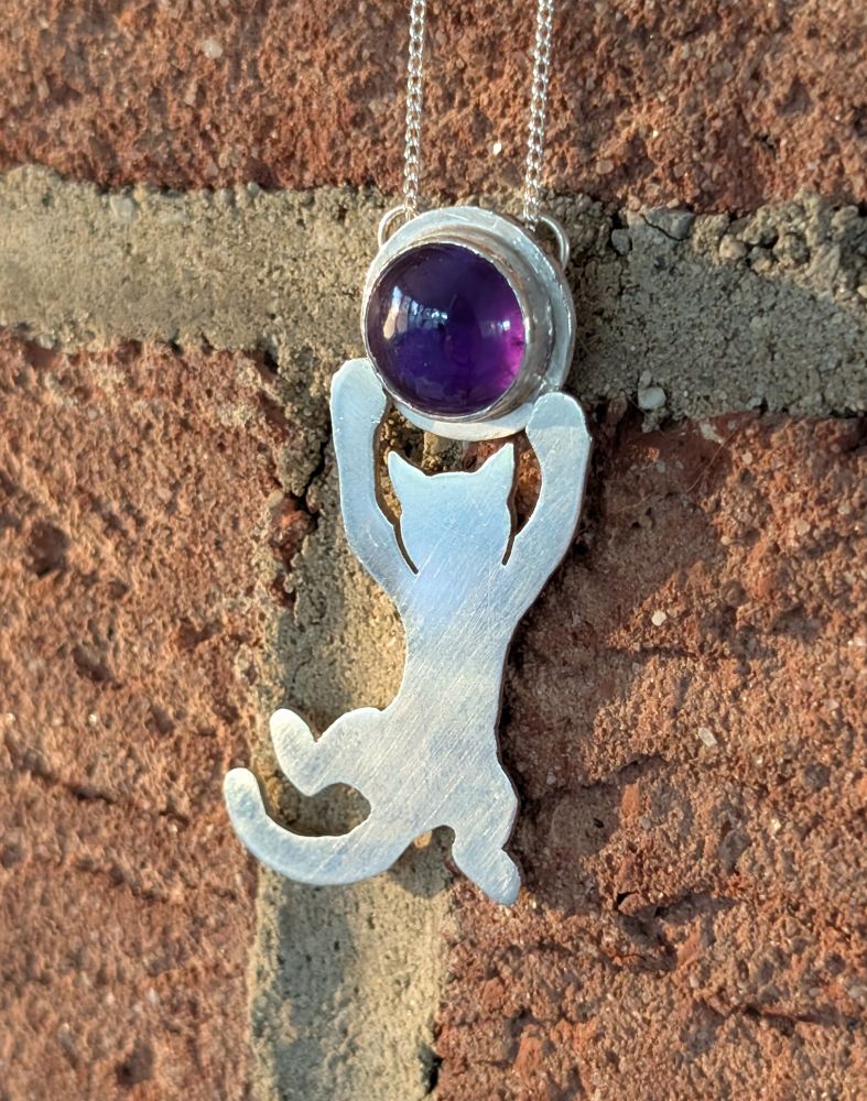 A photo of a silver pendant featuring a round amethyst cabochon, from which the silhouette of a cat is hanging playfully by its paws, as if the amethyst were a ball. It is hanging from a delicate silver chain and displayed against a brick wall 