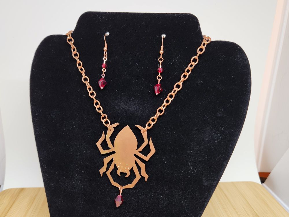 A necklace and earring set in copper with red crystal beads. The necklace is a spider holding a large bead between its front legs. The earrings are long and dangly with a smaller bead in the middle and another, larger one at the end