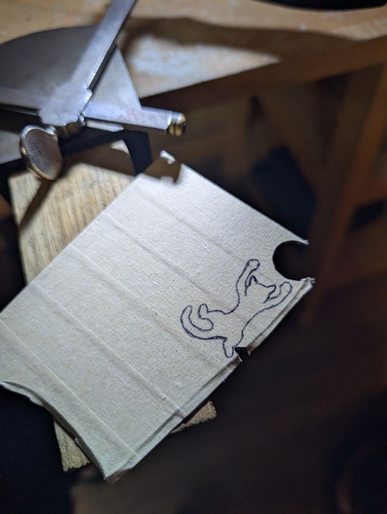A photo of a sheet of silver on my bench. It's covered in masking tape and there is a little cat drawn on it in pen, like it's hanging from something by its front paws. There's a circle already cut out just above the cat.