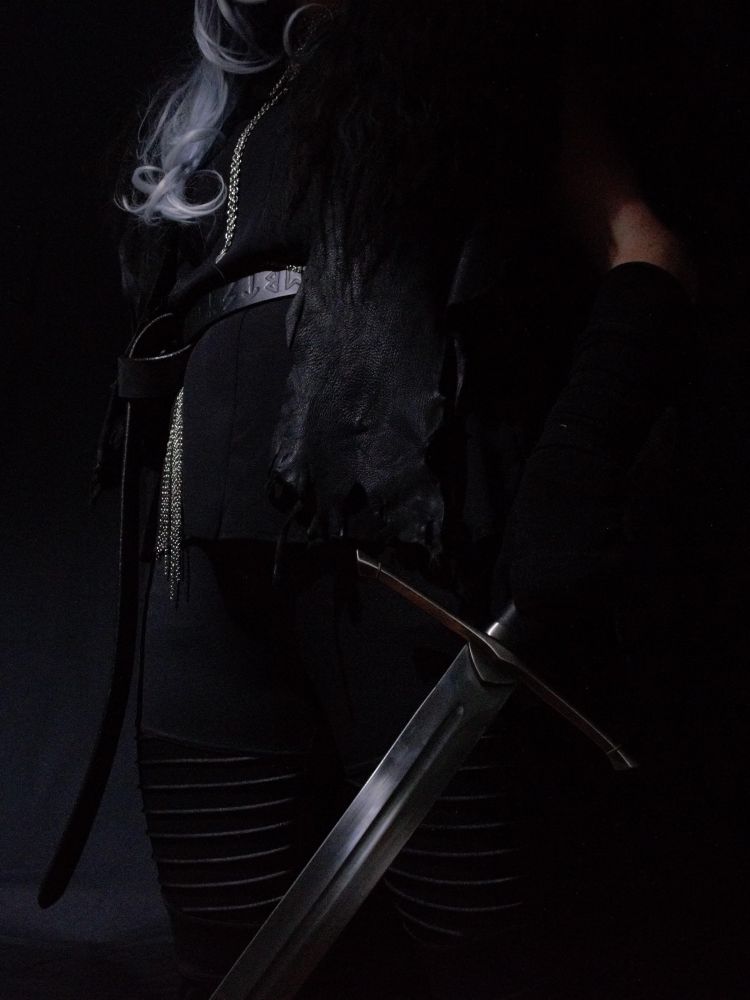 A photo from the same photoshoot as above. It's a shot of me from about the knees up to my neck, standing imposingly as if looking down on the viewer. I am holding my sword in front of me, tip pointing down, and you can see my fancy leggings, belt, and chains hanging from my neck and waist. There's a really nice gleam off the sword that looks really menacing