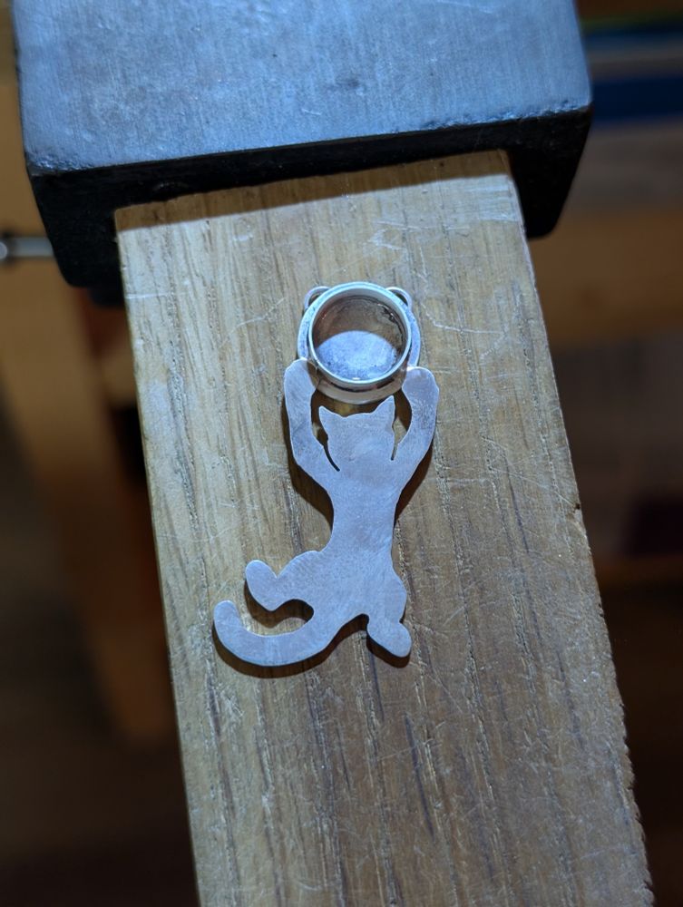 A photo of a silver pendant, mostly finished, sitting on my bench. It shows a cat dangling from its front paws from a circular setting that is, at the moment, empty.
