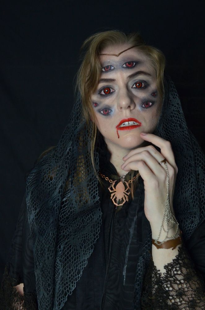 A photo portrait of me in spider-inspired makeup, with six extra eyes painted on my face, red contacts and lipstick, and a drip of blood at the corner of my lips. I am wearing tattered looking, lacy black and grey clothes, with a copper circlet, spider necklace, and spiderweb handflower bracelet. I have one hand held to my face, a curious tilt to my head, as I stare at the camera with my lips slightly parted to show a hint of fangs