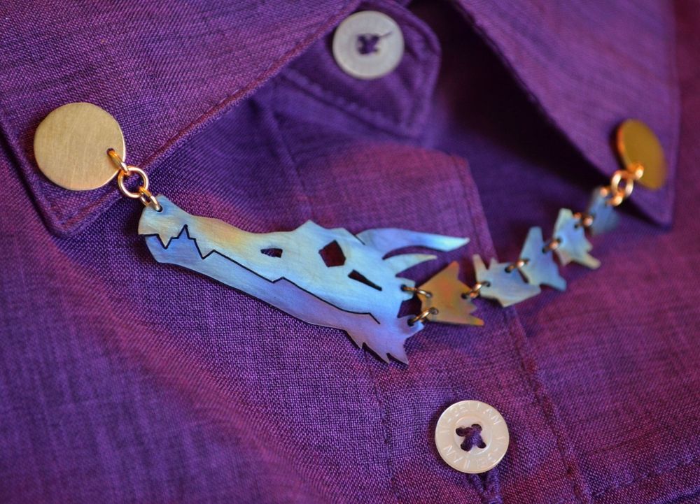 A collar pin in anodized titanium. It is a dragon skull plus several vertebrae, articulated into a chain