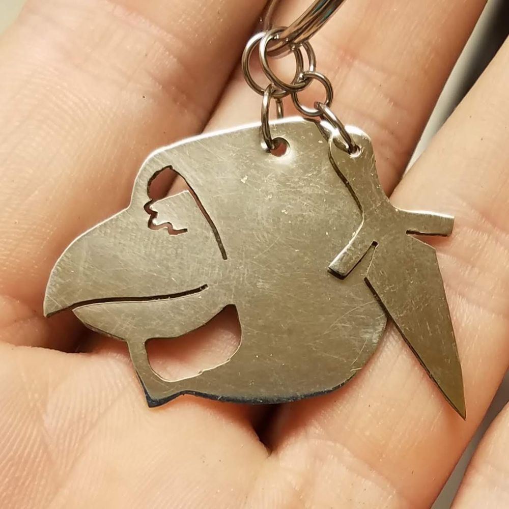 A nickel (silver tone) keychain. It has two parts. One looks like the hooded head of a crow-like person, and the other is a tiny dagger
