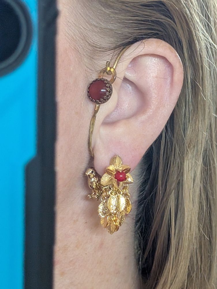 A photo of the ear wraps and earrings described previously being worn. The ear wrap goes all the way around the ear so the red stone sits in front of the tragus. The earring with the leaves gives the effect of the bird charm peeking out from behind foliage, right next to the wearer's neck