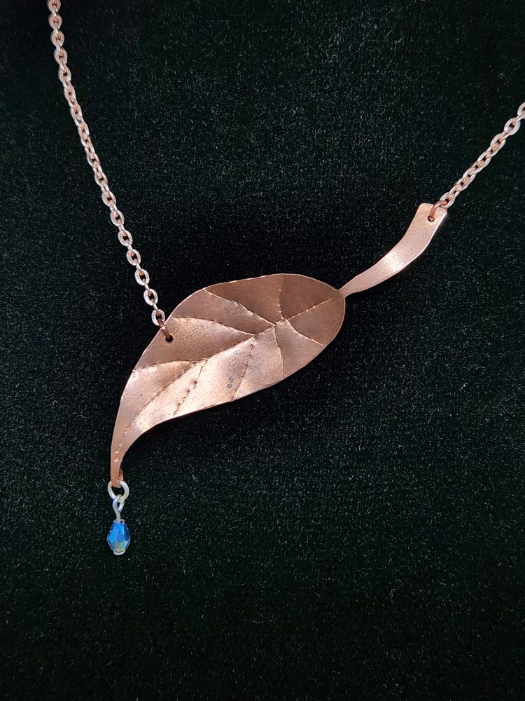 A copper pendant in the shape of a drip-tip leaf, with a blue teardrop bead hanging from the tip like a drop of water. 