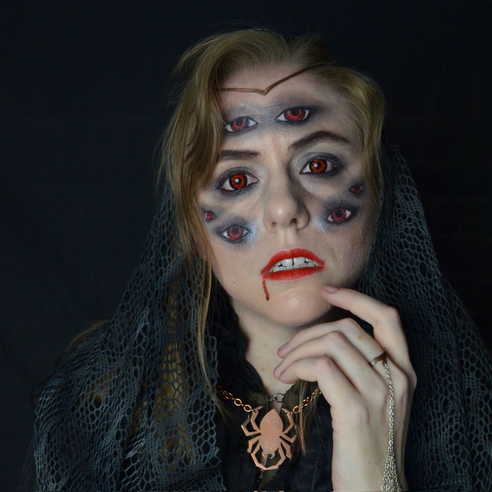 A photo of myself dressed in a lacy black and grey outfit. I'm also wearing a copper circlet, spider necklace, and spiderweb handflower bracelet. I have eyes painted on my face so that I have 8 total, all red because I'm wearing red contacts. I have bright red lipstick and a drip of blood coming from the corner of my eye. One hand is held up to my face and my head is tilted to one side so I look sort of curious as I look directly at the camera.