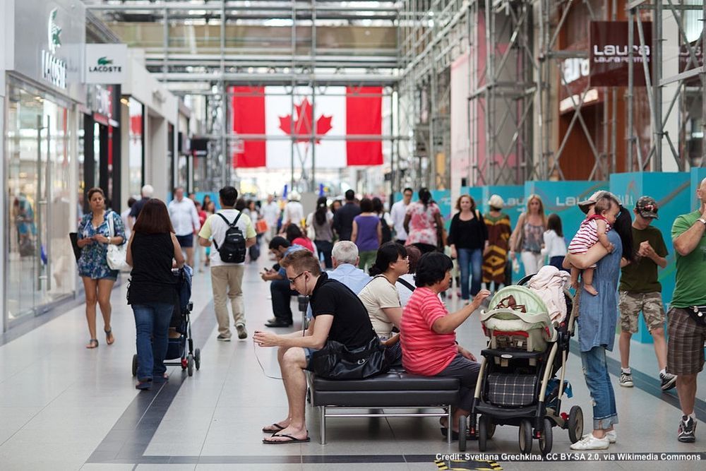 Crowded mall at Eaton's Center in Toronto, with a large Canadian flag in background seen through a large window.