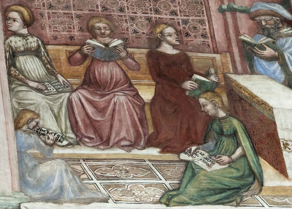 Three young women sit on bench against the back wall with open books. One at the left looks away, distracted by Mary's arrival (not shown in this detail) at the Temple. In the foreground, one young woman dressed in green sits on the floor. She has an open book with visible script and she looks towards a young girl at the right who holds a sheet with the alphabet. To the right, a bearded teacher with a blue robe and hat stands with an open book. The room has richly patterned paneling on the wall and on the pavement.