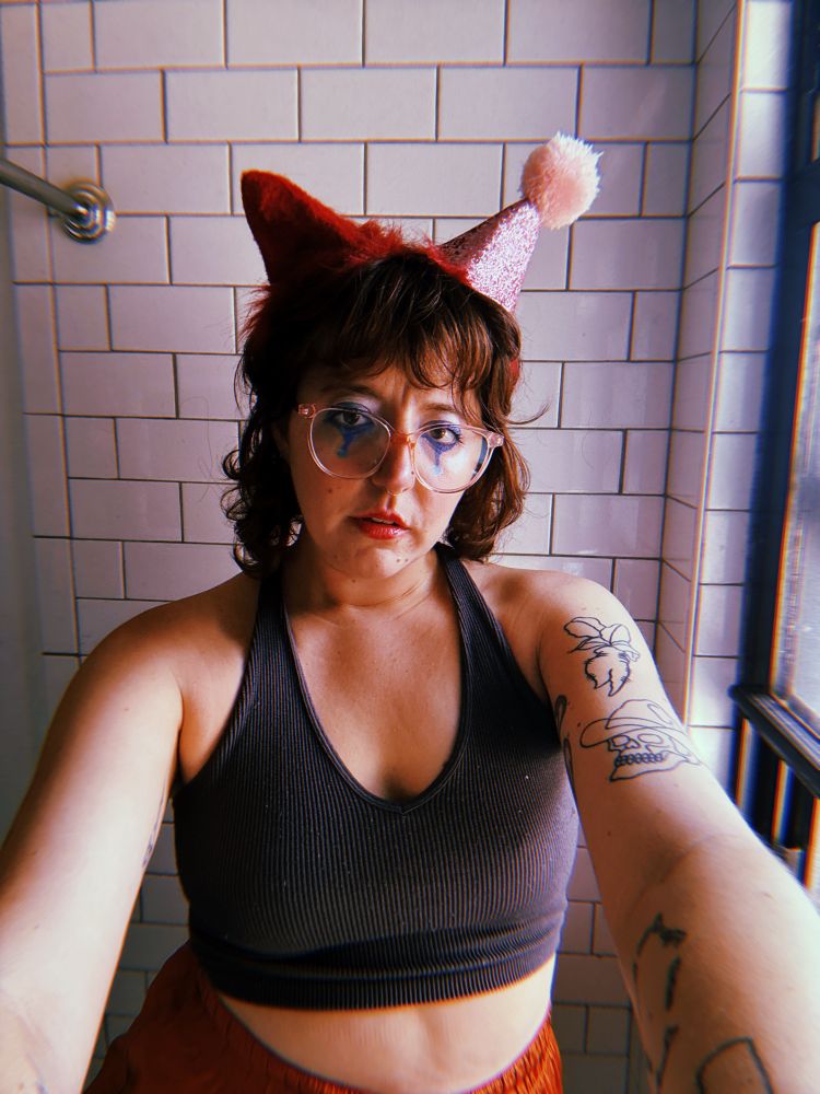 selfie of me, a pale femme, in clown makeup with cat ears and a party hat on as well. i'm standing in my shower. it's dry in here 