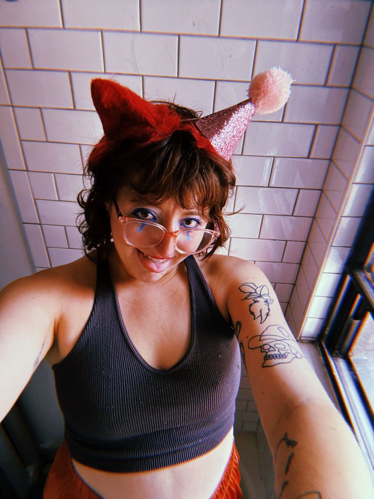 selfie of me, a pale femme, in clown makeup with cat ears and a party hat on, making an evil face at you 