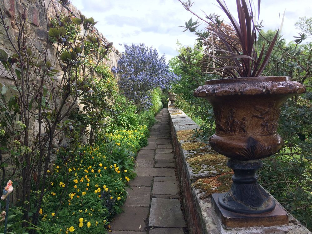 A narrow stone walkway with lots of flowers down one side and a wall down the other side. There's a large stone urn on the wall