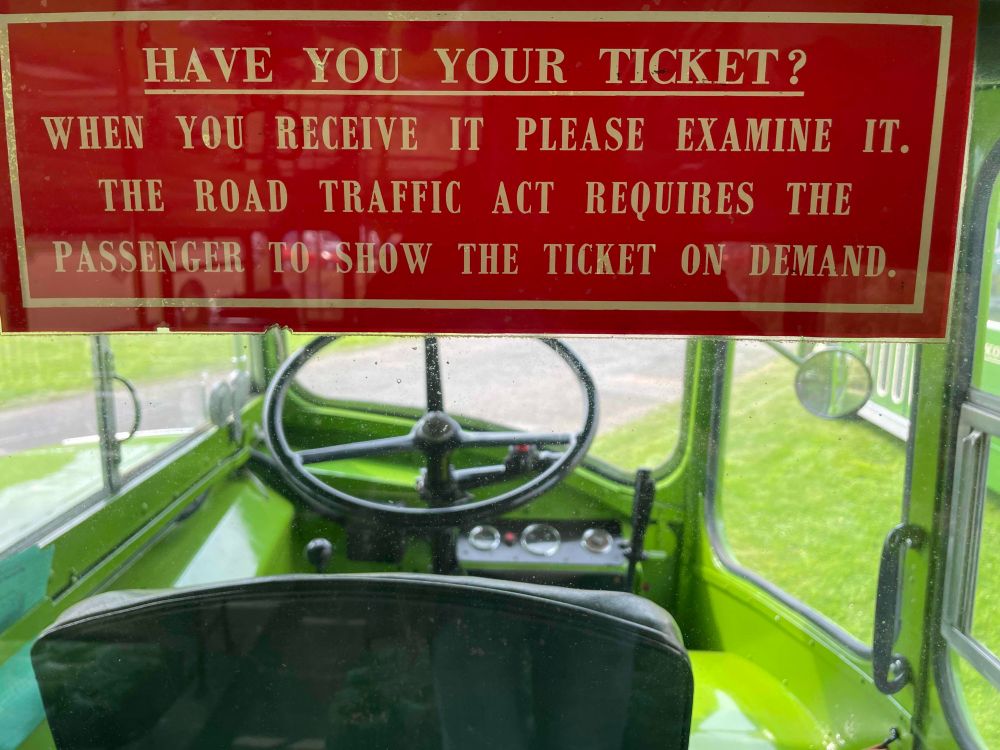 Inside a vintage bus, looking through a window to the driver's cab. There's a sign asking 'have you your ticket?' It goes on to say 'when you receive it please examine it. The road traffic act requires the passenger to who the ticket on demand.'