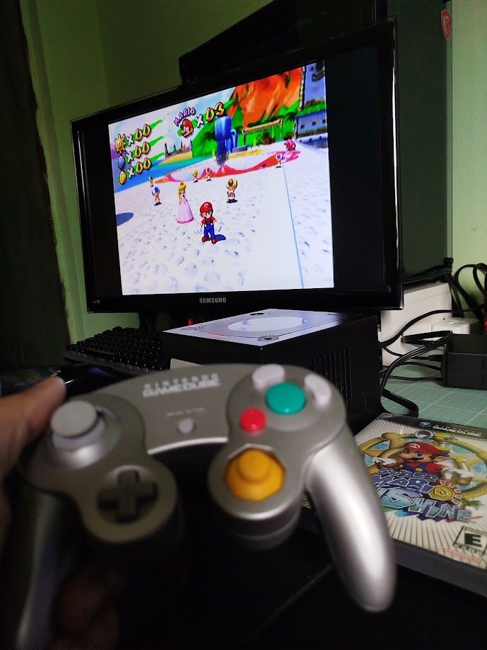 Imagem de um controller de Gamecube e uma Tv com o jogo Super Mario Sunshine rodando, no geral uma bancada com a TV, o gamecube, e a capinha do jogo.