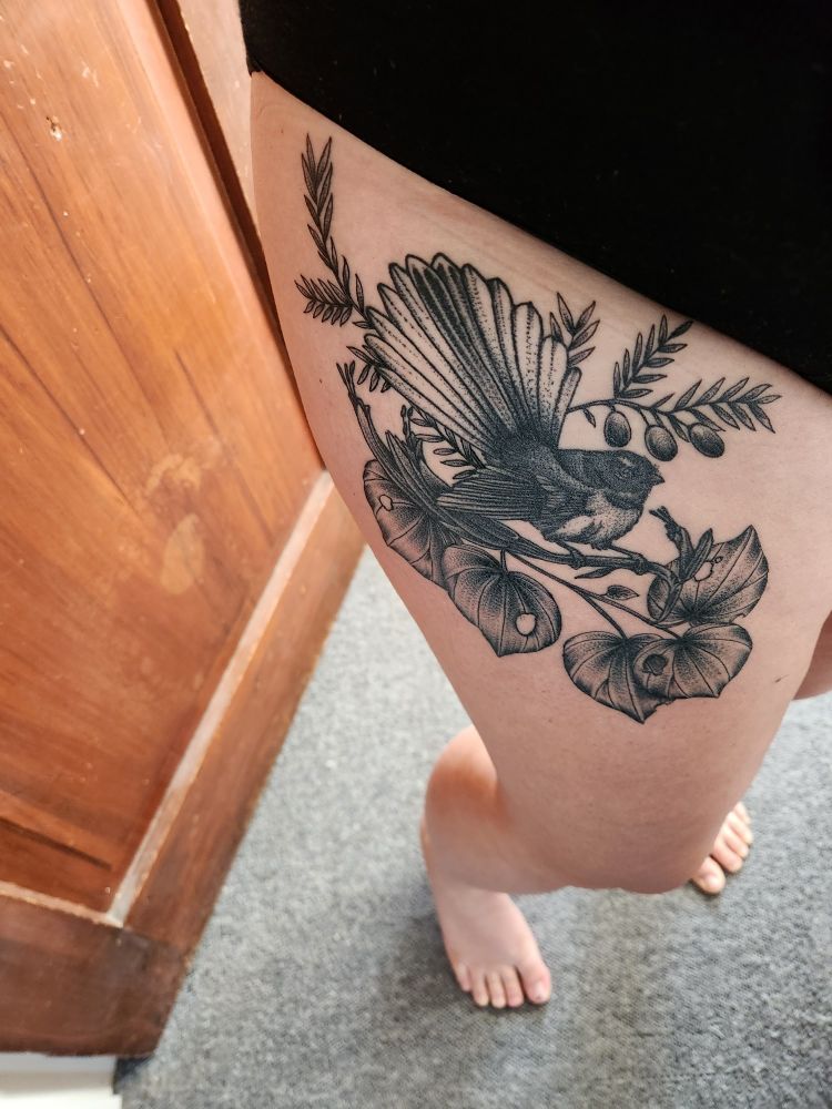 Image shows a tattoo at the top of Tori's thigh. The tattoo is a pīwakawaka/fantail with its tail feathers fanned out and an angry expression on its face. It is perched on the stem of a harakeke flower, and surrounded by kawakawa leaves below and miro berries & stems above