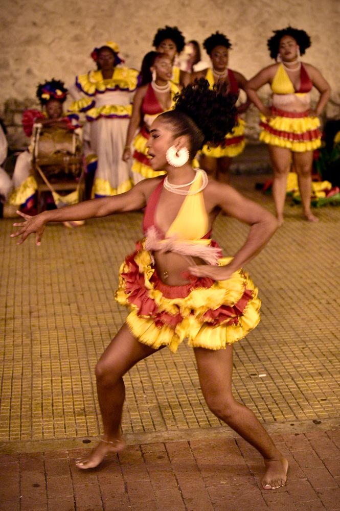 A female dancer in the foreground, with red and yellow skirt and top. In the background other dancers admire her.