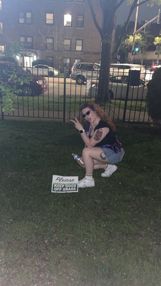 Making a face and kneeling in the grass by a sign that reads “please keep dogs off grass”