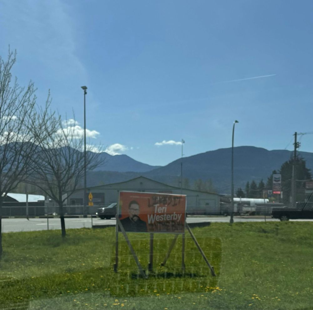 Political campaign sign for NDP candidate Teri Westerby on a grassy area in Chilliwack, BC, with mountains and buildings in the background. The sign is spray-painted with black graffiti.