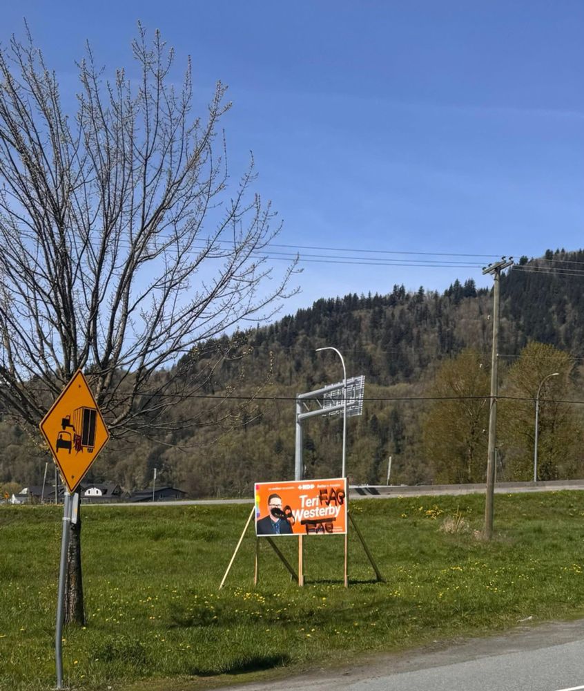 Campaign sign for NDP candidate Teri Westerby positioned near a highway and tree in Chilliwack, BC. The sign is heavily vandalized with the homophobic slur “FAG” spray-painted over both the candidate’s face and name in bold black letters. A yellow caution sign for truck traffic is visible nearby.