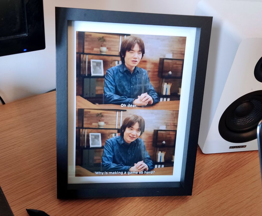 A framed print of two screengrabs from the last Kirby Air Riders Direct. Game Director Masahiro Sakurai is saying 'Oh Dear', 'Why is making a game so hard?'. The framed picture is sitting on my desk.
