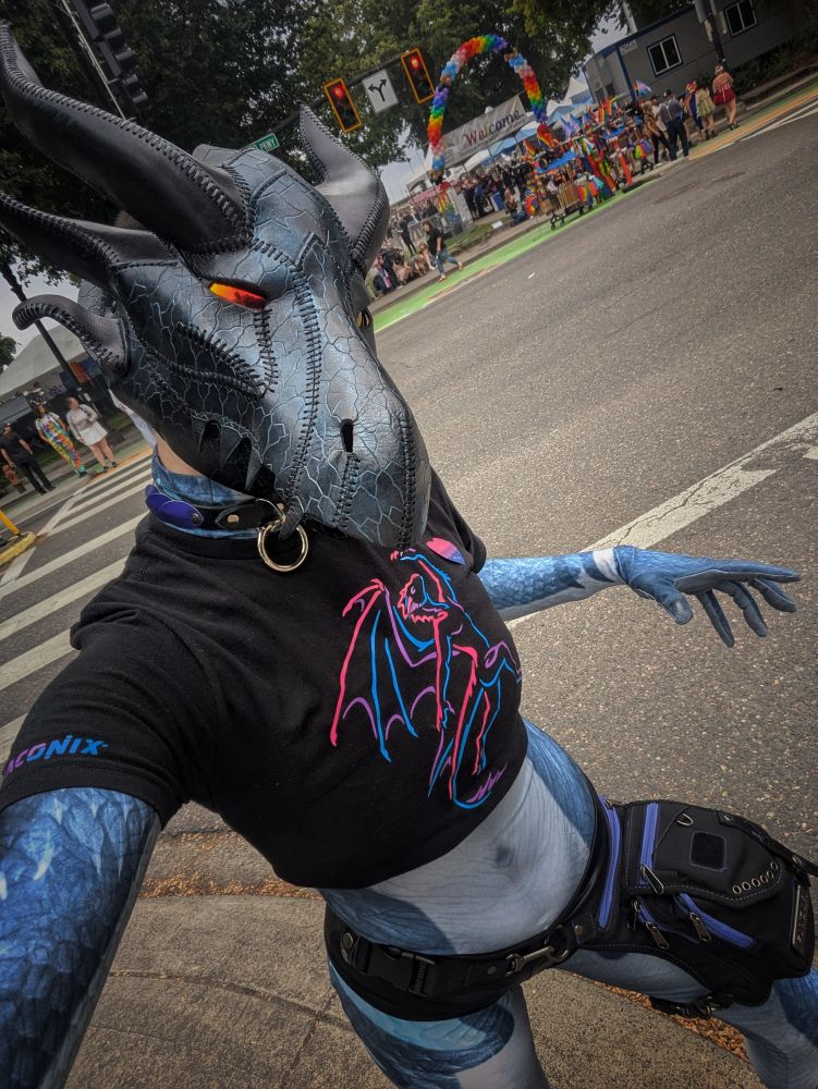 Selfie of Lycosaurus at Portland Pride 2025. Wearing a leather dragon mask, scaly patterned spandex petsuit (bodysuit), & crop top featuring art by andraconix of a dragon in bisexual/androgyne pride colors