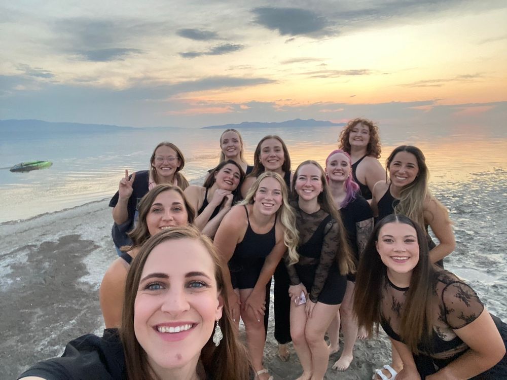 12 women dressed in black smile at the camera while standing on the shore of Utah Lake. The sun is setting behind them casting a orange and yellow hue over the clouds and sky. 