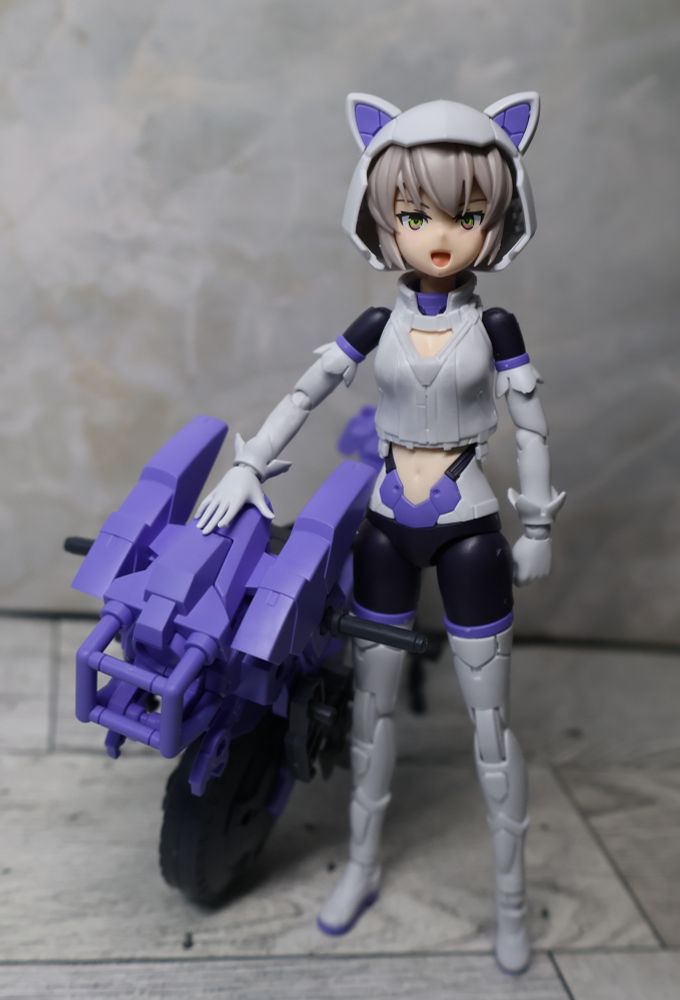 A small plastic girl with an open mouth and cat-ear helmet stands beside a small plastic mono-cycle. She is wearing a white crop top and  black leggings with white thigh-high boots. One hand rests on the purple motorcycle.
