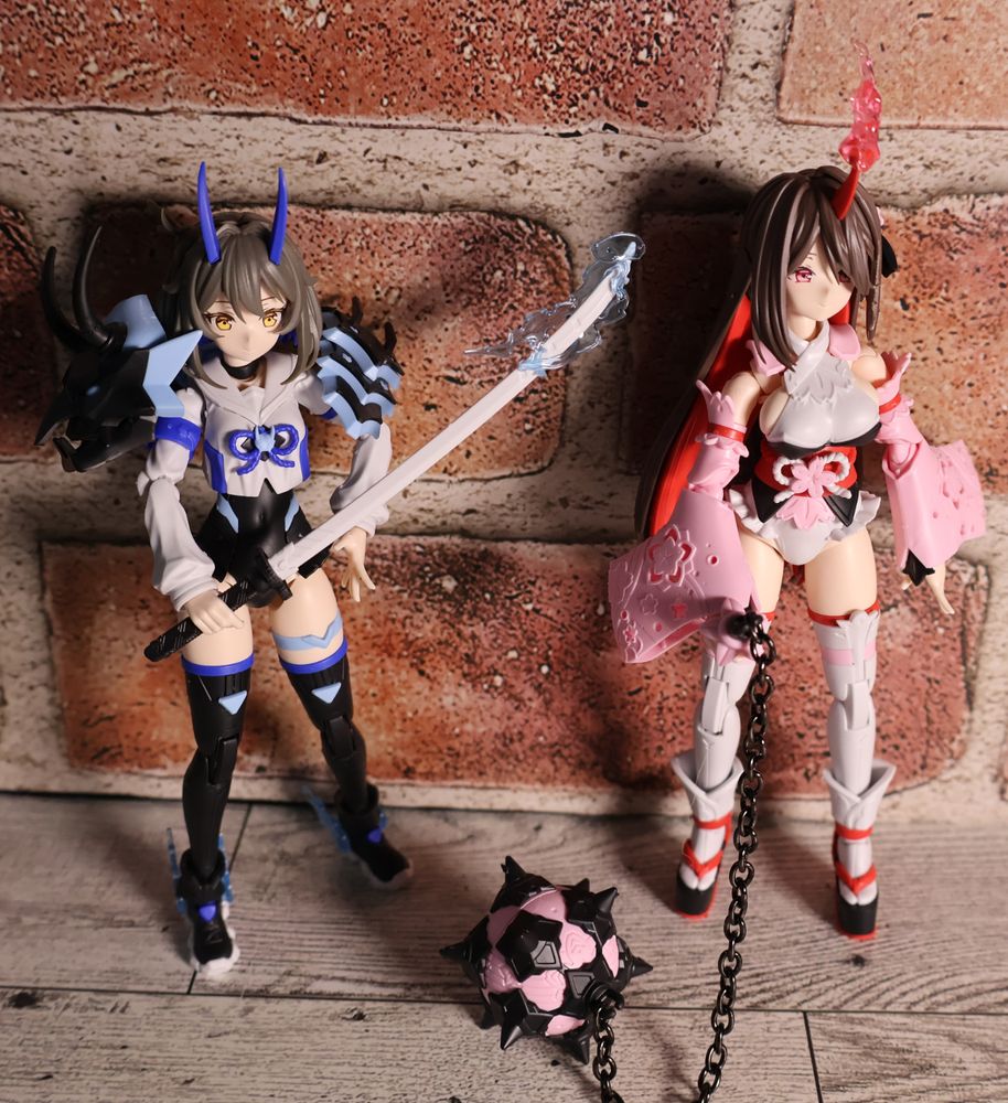 A pair of plastic model girls pose together against a brick wall on a wooden floor. The left model is done in shades of blue and black. She holds a long sword with pale blue lightning ringing the blade. A large demon-head stole wraps around her shoulders, and blue demon horns poke up through her hair. On the right is a slightly taller figure (taller because of her shoes) with a red, flame-wreathed horn on her head. Her outfit is shades of pink, red, and white, with a beribboned sash around her waist and flowing pink sleeves. She holds the handle of a meteor hammer, which rests on its spikes beside her. 