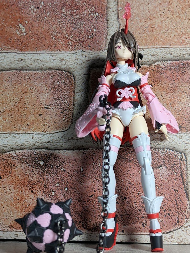 A small plastic girl with a red, flame-wreathed horn on her head. Her long brown hair frames her face. Her outfit is shades of pink, red, and white, with a beribboned sash around her waist and flowing pink sleeves. She holds the handle of a morning star (yes, this is how it's referred to in her descriptions, although the longer chain suggested meteor hammer), which rests on its spikes beside her. 