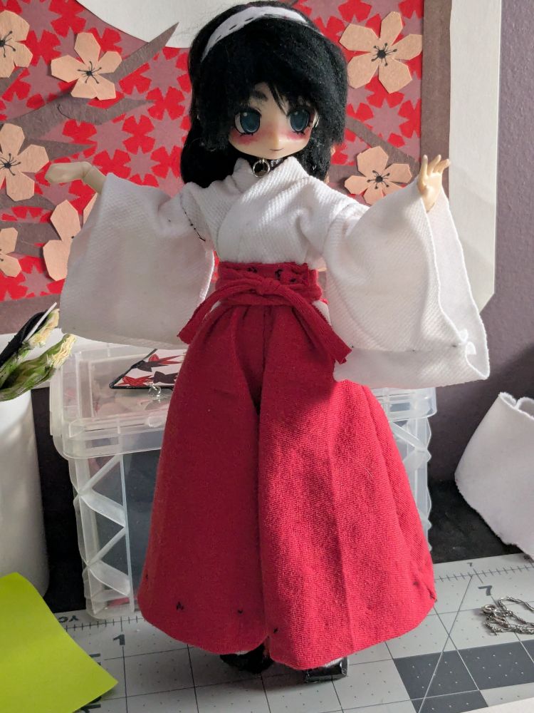 photo of a doll wearing a white kimono and red hakama pants. she is standing in front of a paper flower backdrop on a desk