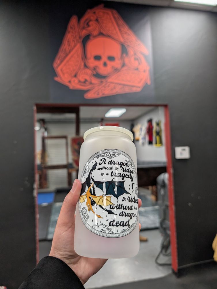 A picture of a frosted glass tumbler with a Fourth Wing circular design of 2 dragons, one large black one and one smaller gold one, and the quote "A dragon without its rider is a tragedy. A rider without their dragon is dead" on it.