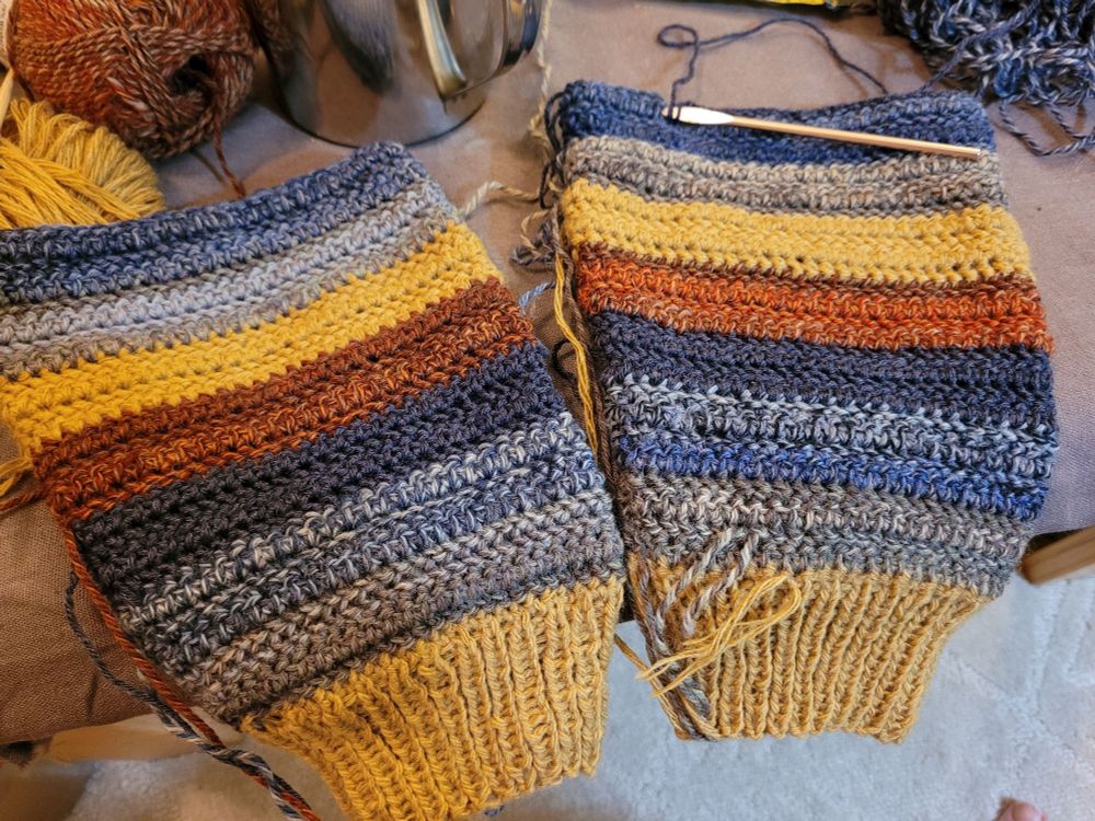 Photo looking down at the start of two crocheted sweater sleeves, about 9 inches long each, with a tan colored cuff and alternating stripes in earthy greens, blues, tan, and red.
