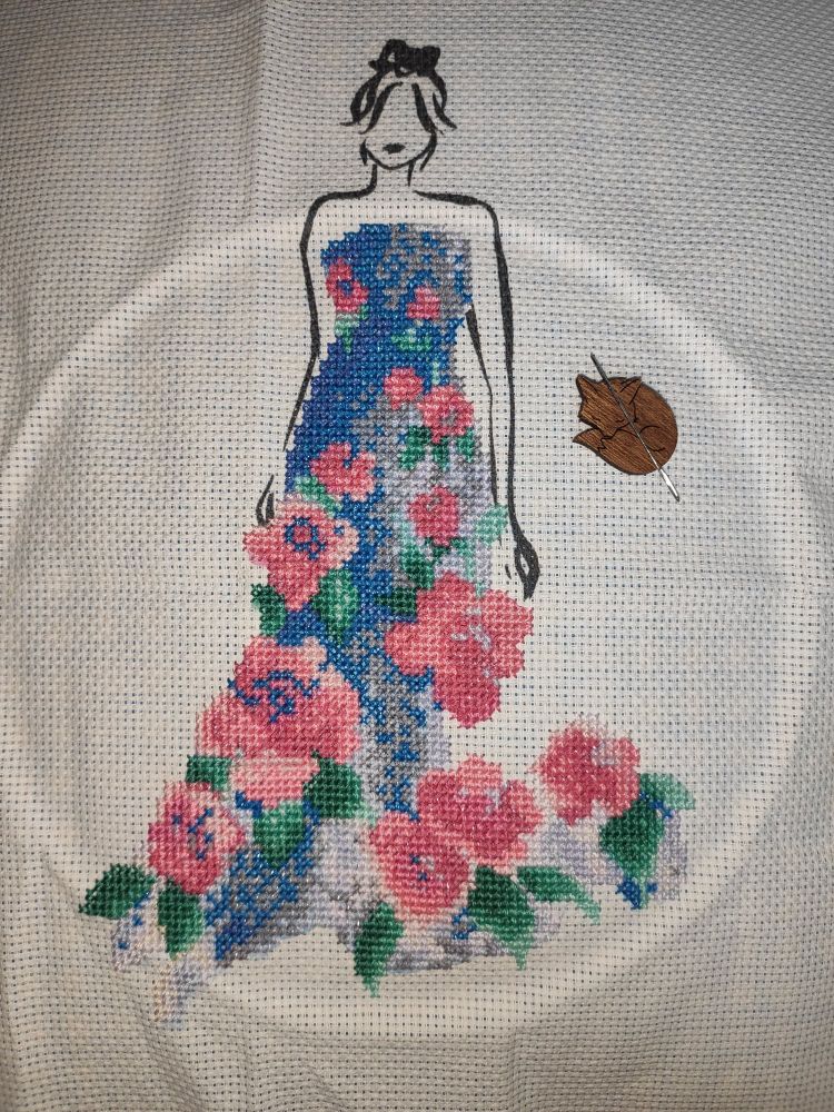 White Aida fabric with a black silhouette of a woman's head and arms printed on the fabric. Her dress (cross-stitched) is blue and silver with pink roses and green leaves, the flowers popping outwards. Some of the threads have a silvery sheen to them.