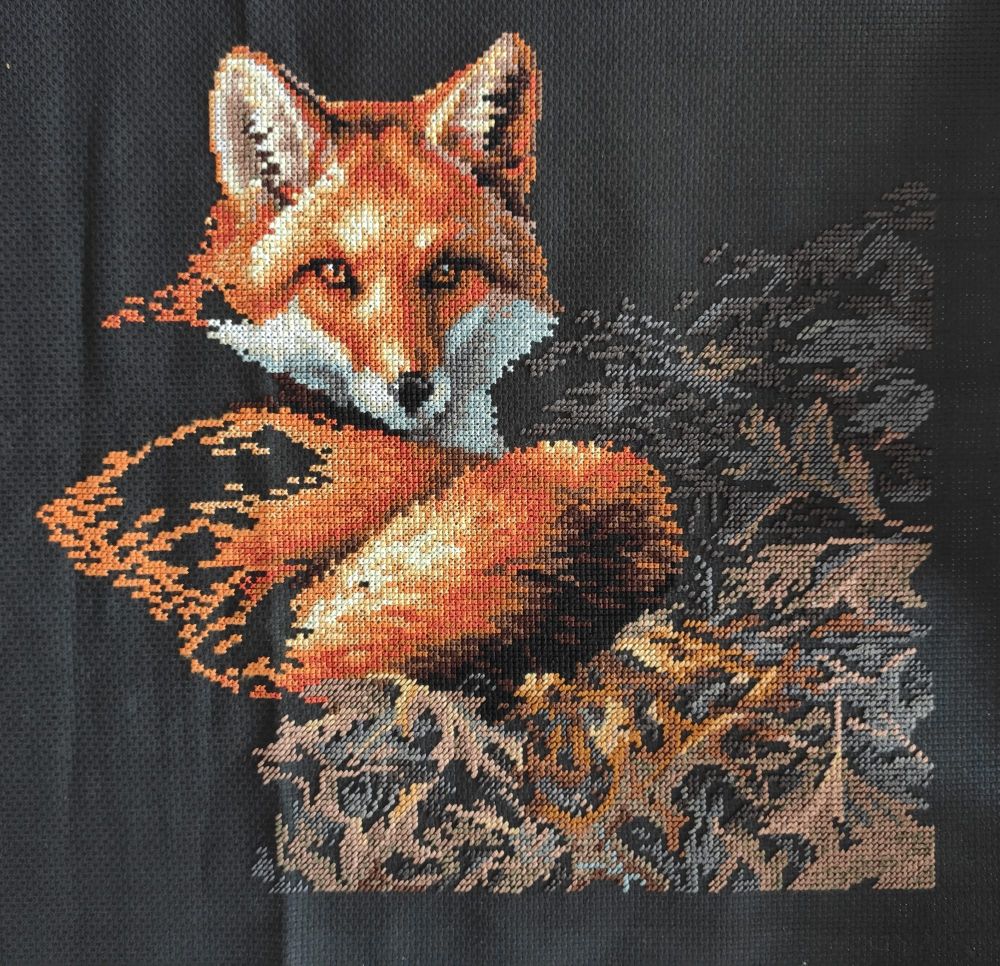 A partially completed cross-stitch of a fox curled up on a bed of leaves on black Aida. The leaves are half-crosses in shades of brown and grey, while the fox is in shades of orange, yellow, red and white.