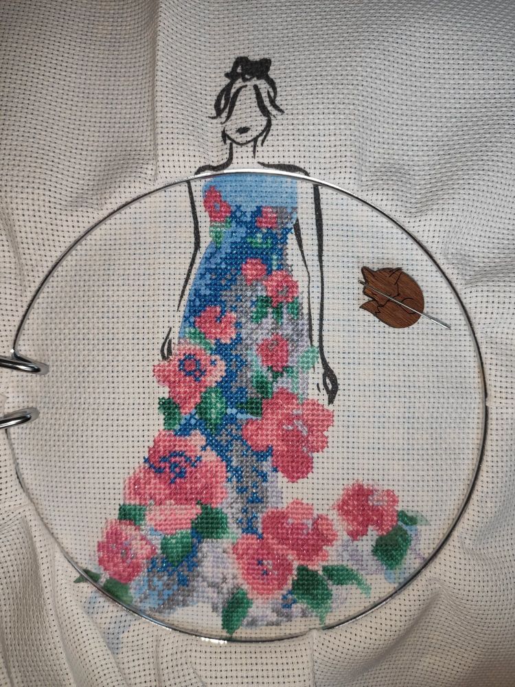 A counted cross-stitch pattern of a woman wearing a blue and silver dress with pink roses that pop out. Some of the threads are shiny. The woman is a silhouette outlined in black on white Aida. Most of the cross-stitch is done.