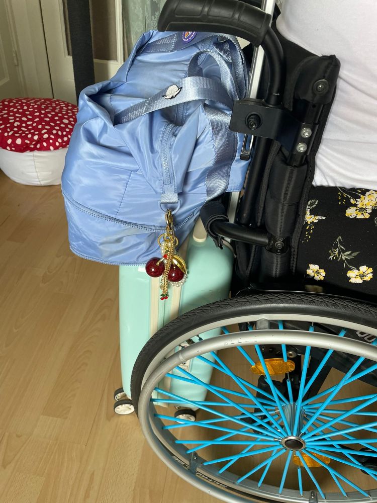 Een foto van Marloes' rolstoel, met blauwe spaakrietjes. Een rolkoffer en een grote schoudertas zitten op onzichtbare wijze aan elkaar en de rolstoel vast. 