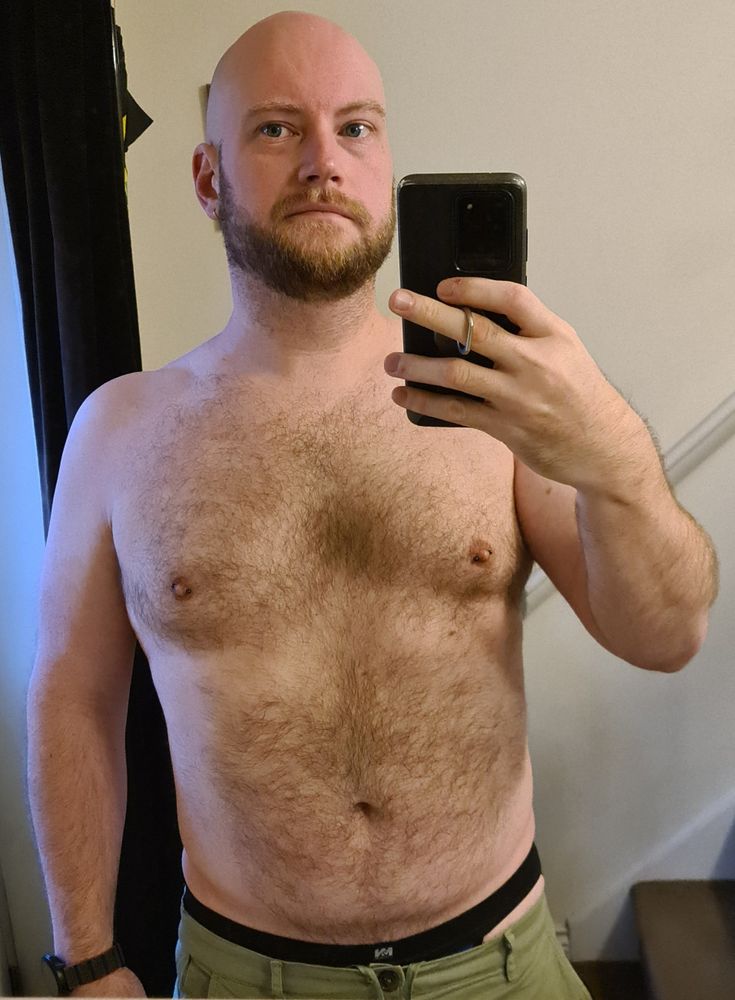 A picture of me, a bald and bearded white cis guy, standing in front of a mirror. I'm shirtless and have light brown hair on my chest and stomach. The waistband of my jock is visible as my green chinos have sagged down my waist.