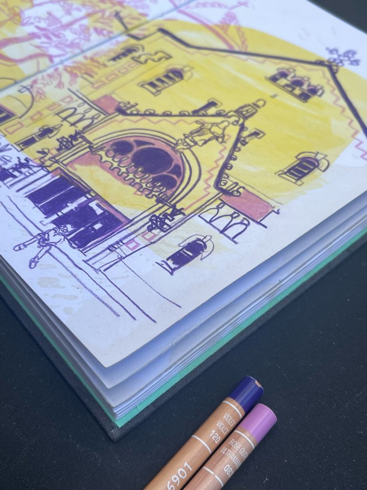 This is the sketchbook from a different angle. You can barely see some cream coloured stains on the page and on the side of the sketchbook. 2 coloured pens are laying in front of the open book.