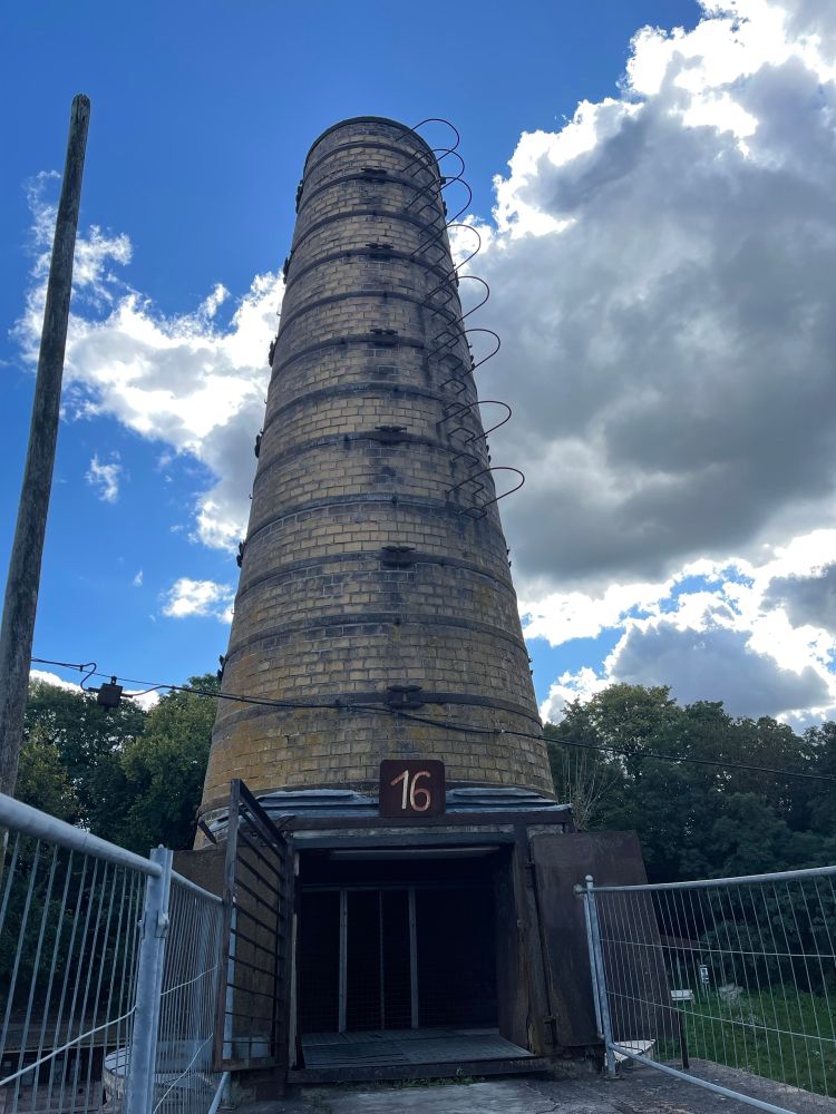 A chimney with an open door at the bottom. There’s a rusty sign with a 16 on top of the door.