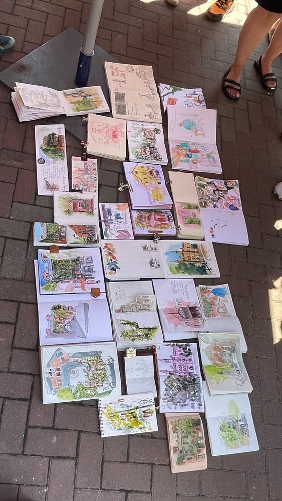 A big throwdown of sketchbooks on the floor. Open sketchbooks in the middle , people stand around, so some feet are visible.