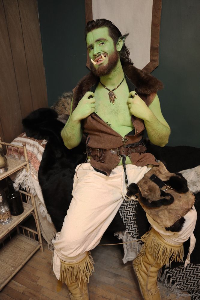 a green orc man sitting on a bed, holding open his brown doublet exposing his green chest. Wearing tan pants, and moose hide mukluks. He's looking at you with a smile showing off his tusks.