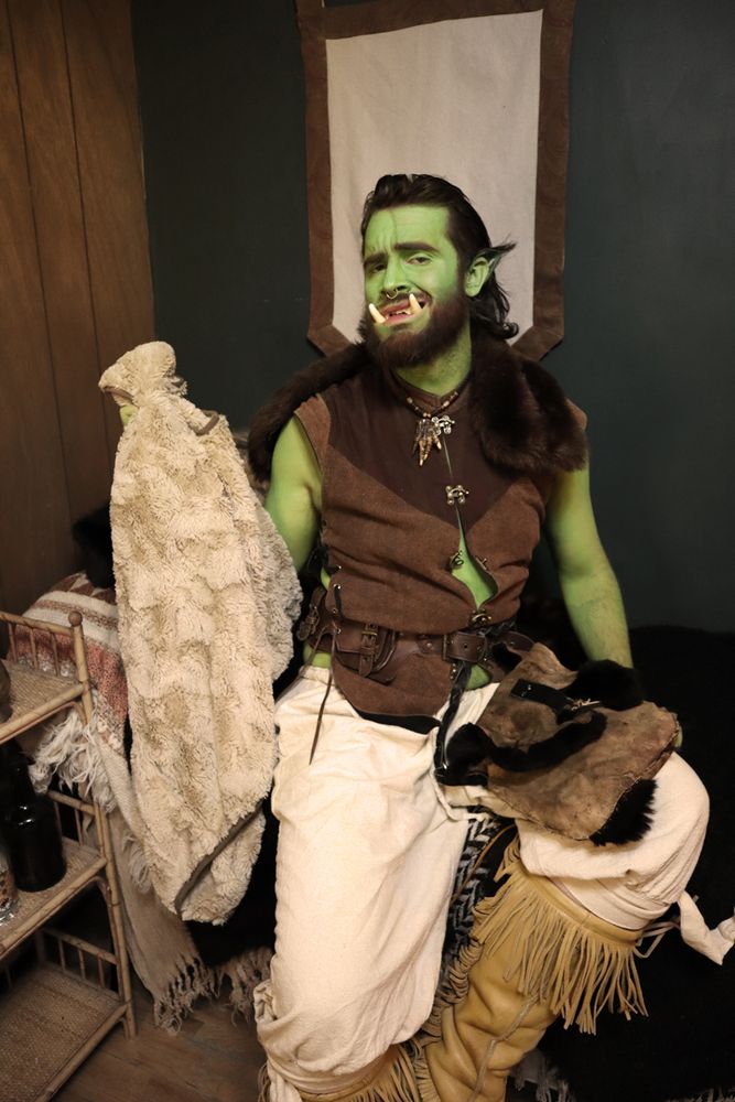 a green orc man sitting on a bed, holding a tan furred shawl, wearing a brown doublet, tan pants, and moose hide mukluks. He's looking at you with a smile showing off his tusks.