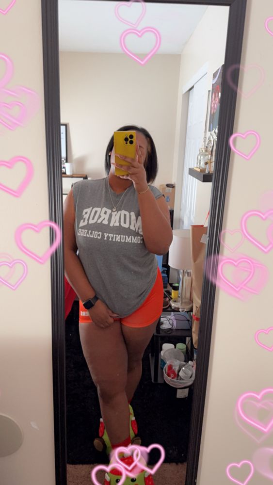 A brown skinned woman wearing a college grey t-shirt and orange boxers taking a picture, standing in a bedroom with her legs crossed in front of a mirror