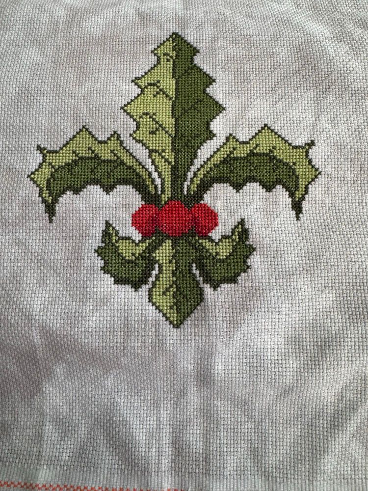 Cross stitched Fleur de lis made of green holly and three red berries on grey fabric 