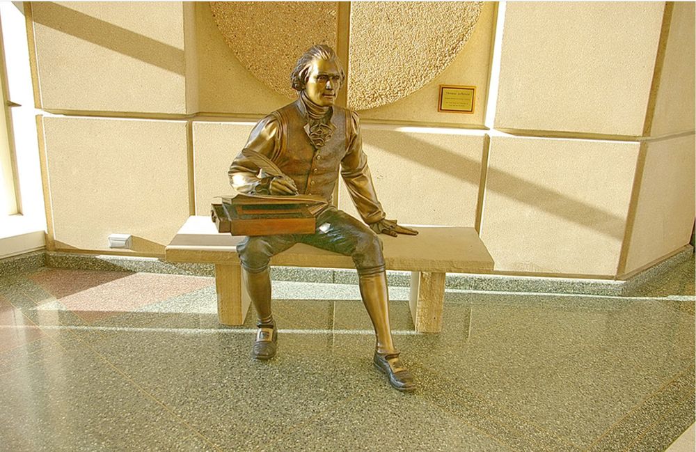 Sculpture of Thomas Jefferson, sitting on bench with portable writing desk on his lap. He's writing with a quill pen.