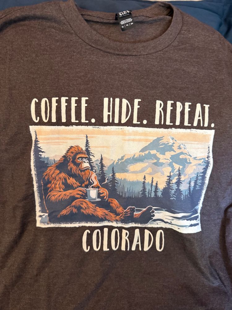 Brown T-shirt with a picture of Sasquatch drinking a cup of coffee, the Rocky Mountains behind him. Words above “Coffee. Hide. Repeat. Word below “Colorado"
